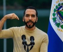 Fotografía de Bukele apuntándose así mismo con una remera beige. De fondo la bandera de El Salvador